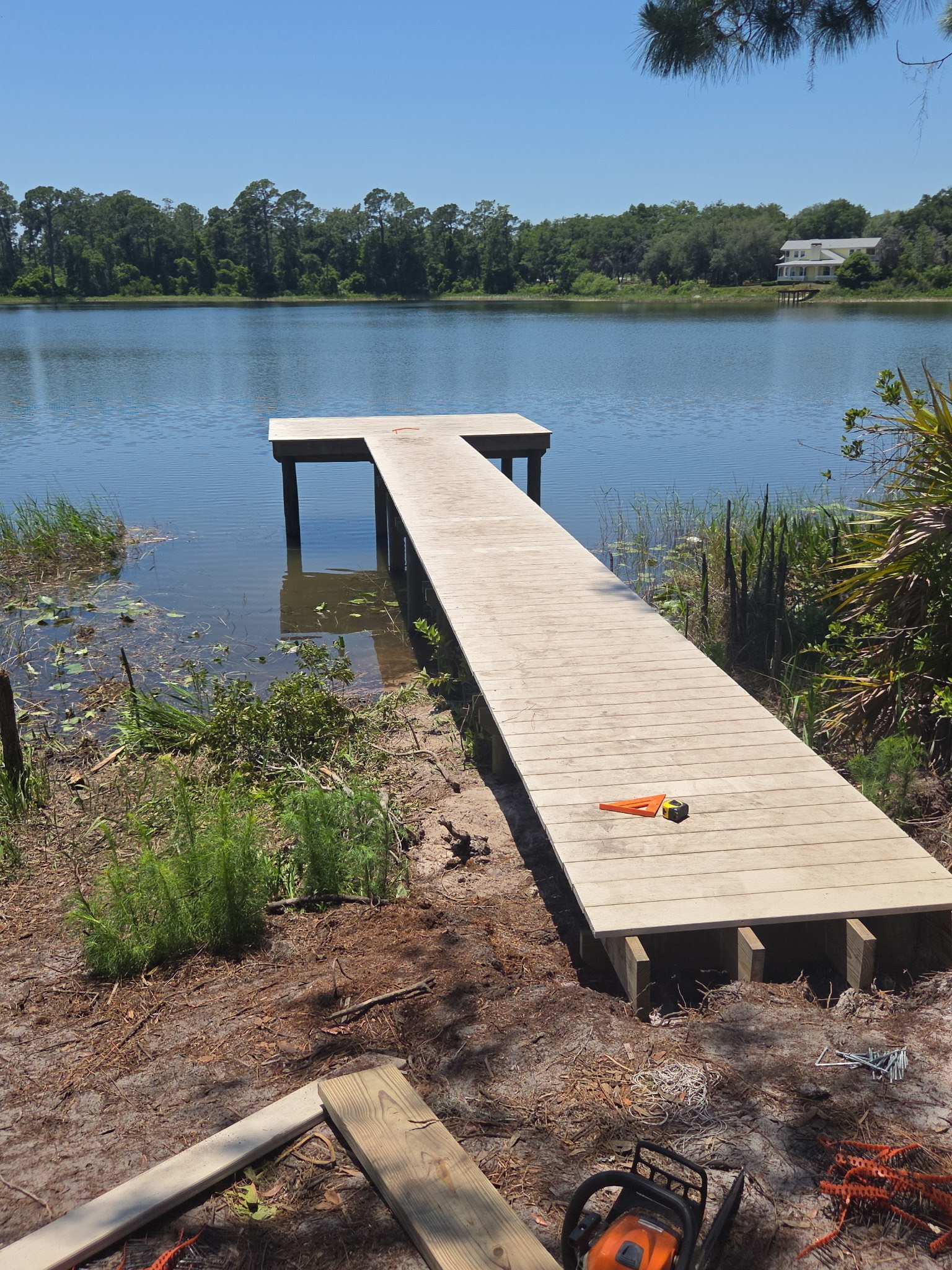 1722061785807679698 - JSC Contracting Inc Unfinished wooden dock extends into a calm lake, with lumber and tools scattered on the muddy shore nearby.