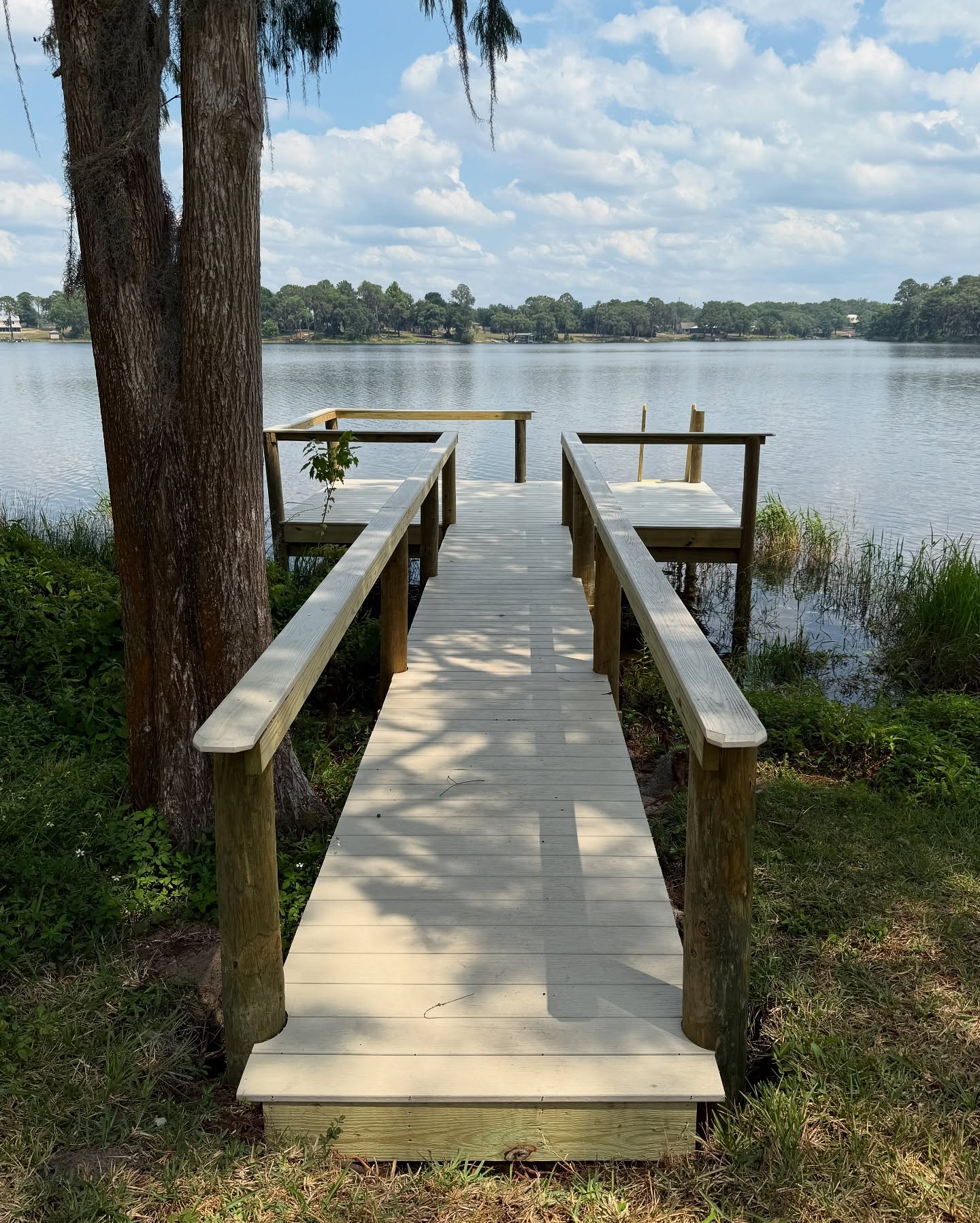 494402360_17927680173049257_6599222707276575717_n - JSC Contracting Inc Wooden dock extending over a calm lake, with grassy banks and trees, under a partly cloudy sky.