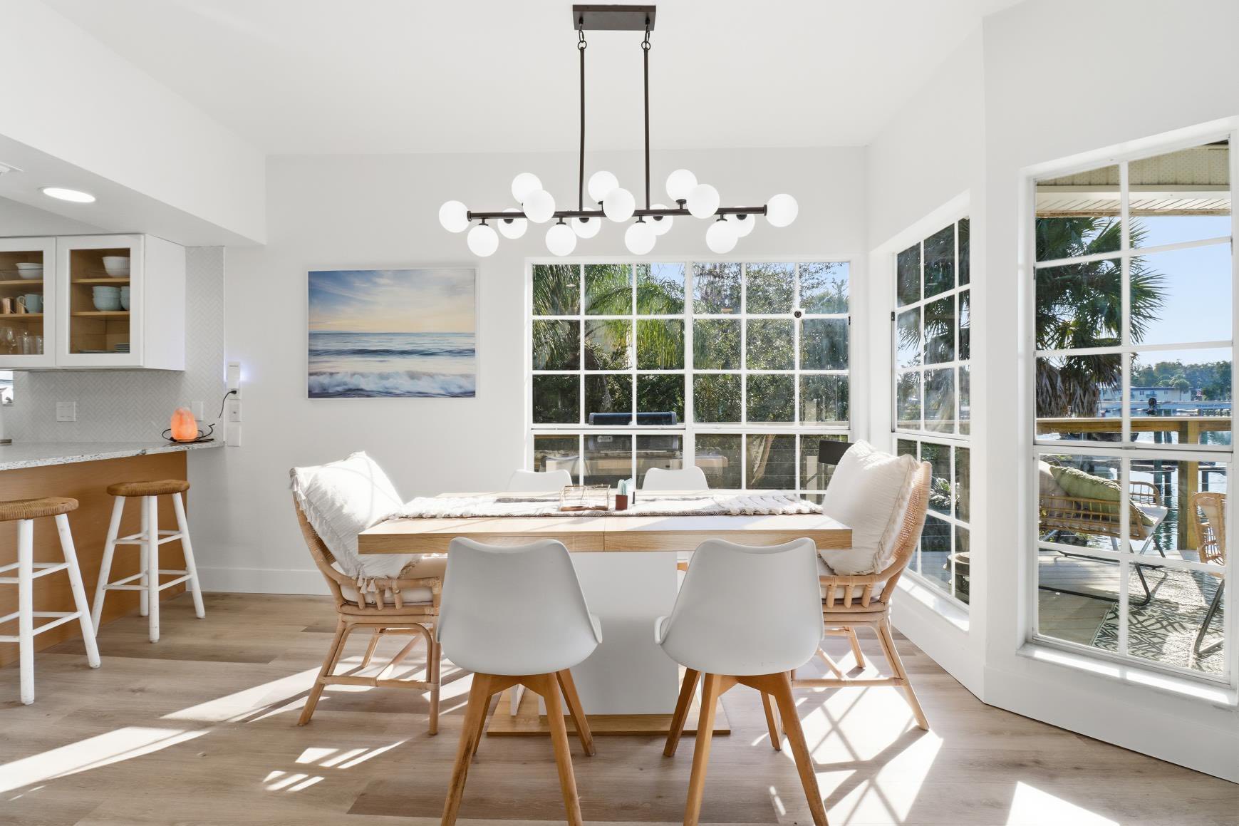 Bright open dining area with a wooden table and white chairs, and a modern globe chandelier hung over the space near large windows overlooking palm trees outside.