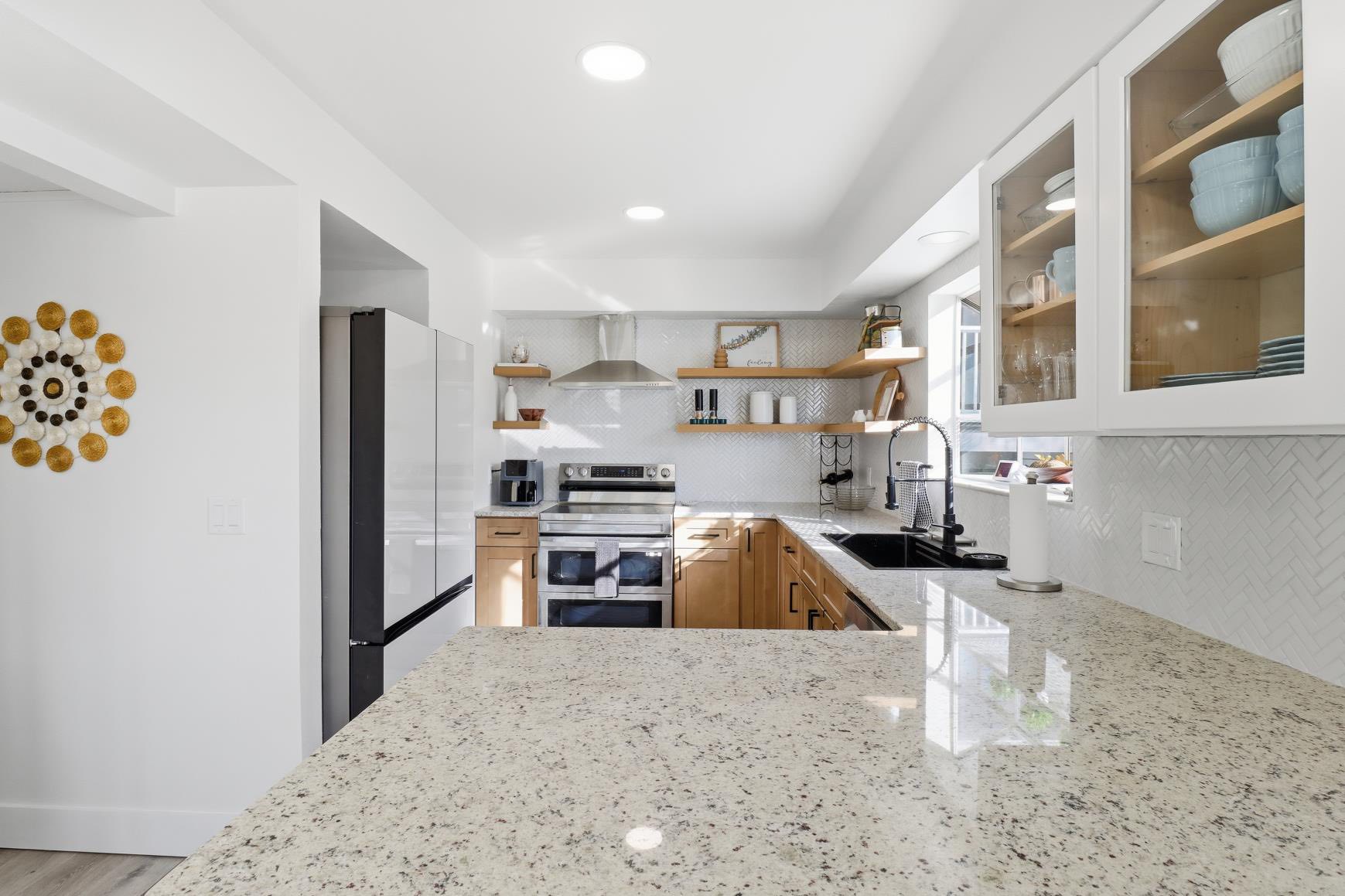 Bright modern kitchen with a speckled granite island in the foreground, white cabinets, stainless steel range, and open wooden shelves.