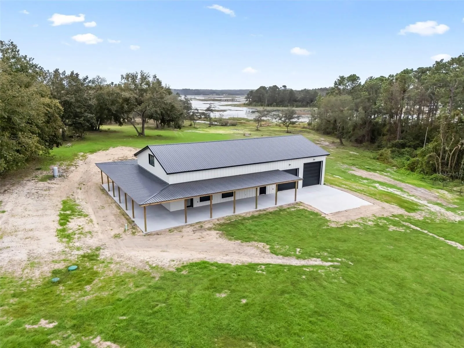 Aerial view of a modern white metal-roofed building with a long covered porch on a green, open field near a lake and surrounding trees.
