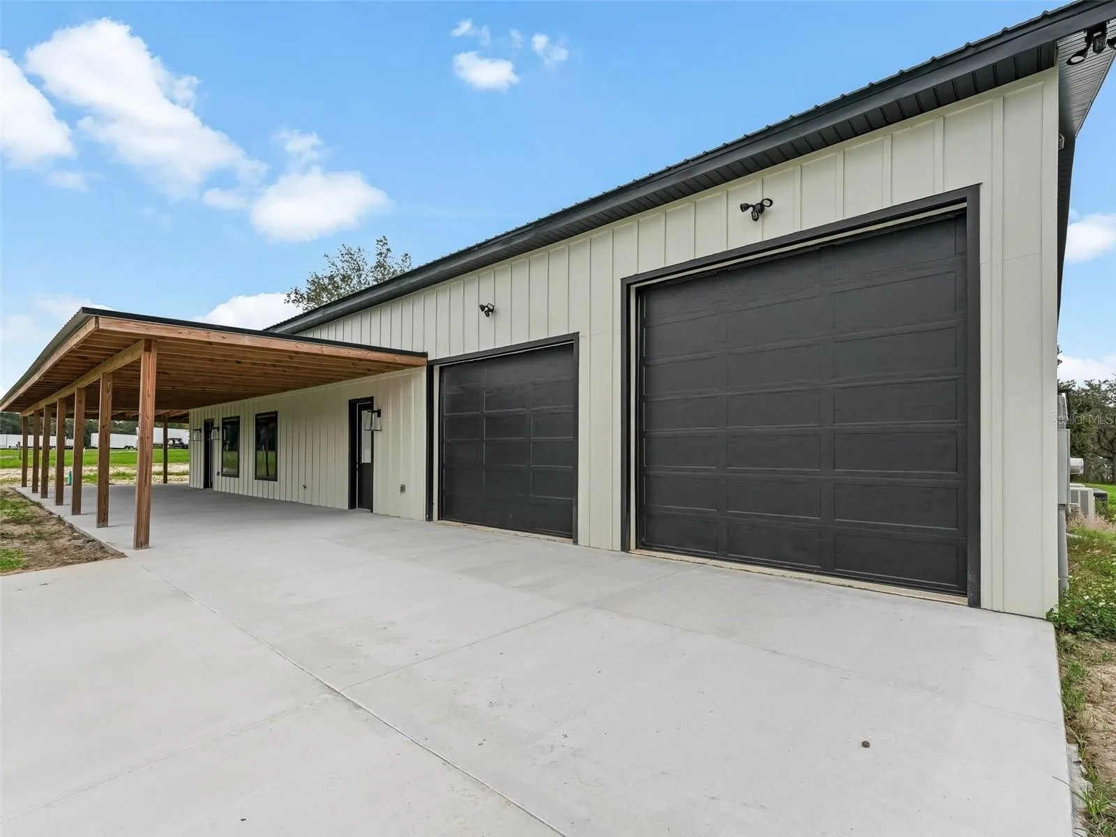 Industrial-style building with two large black roll-up garage doors under a wooden carport on a concrete lot