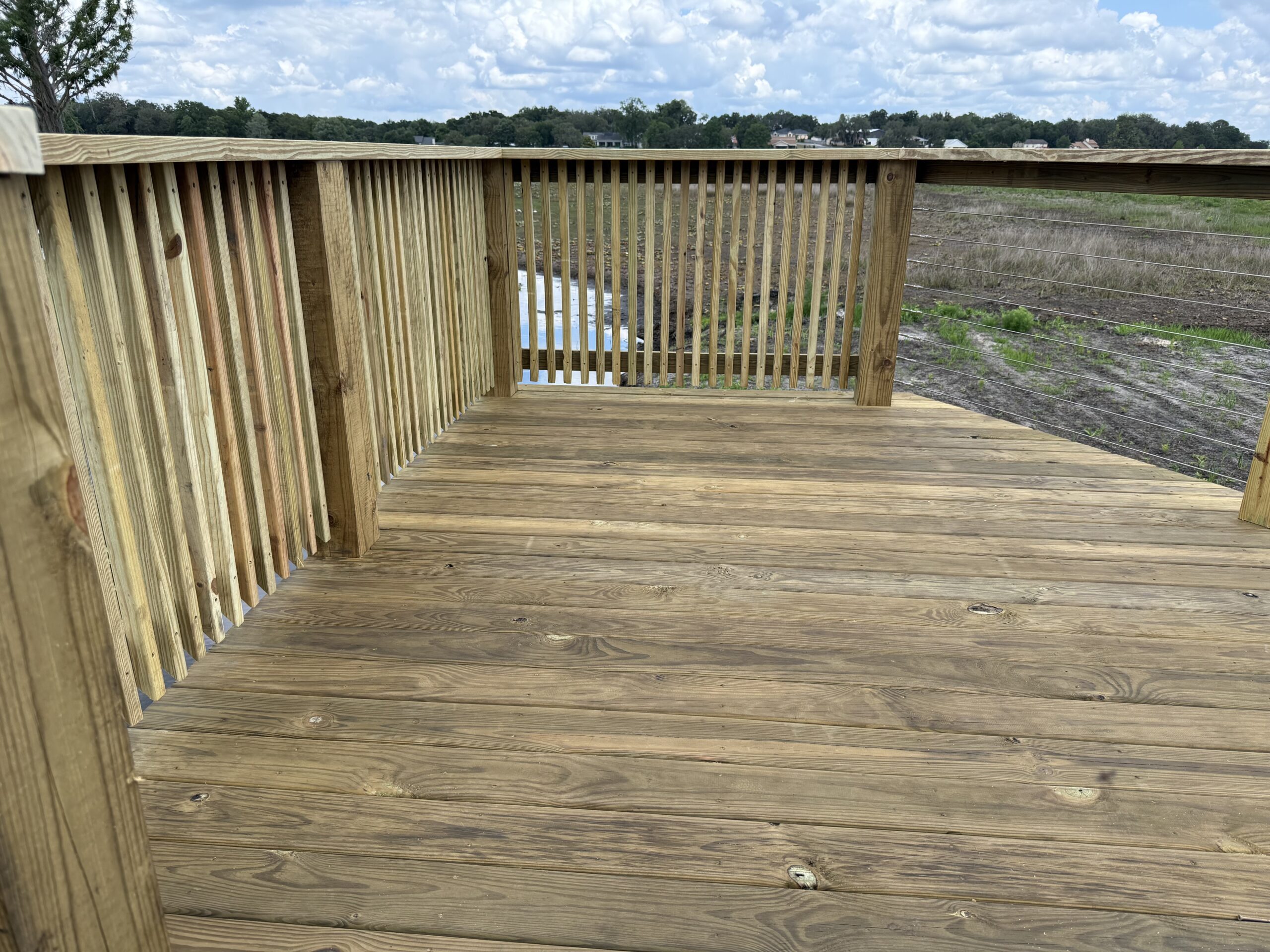 IMG_2567 - JSC Contracting Inc Wooden deck railing and floor overlooking a grassy field with distant houses under a cloudy sky.