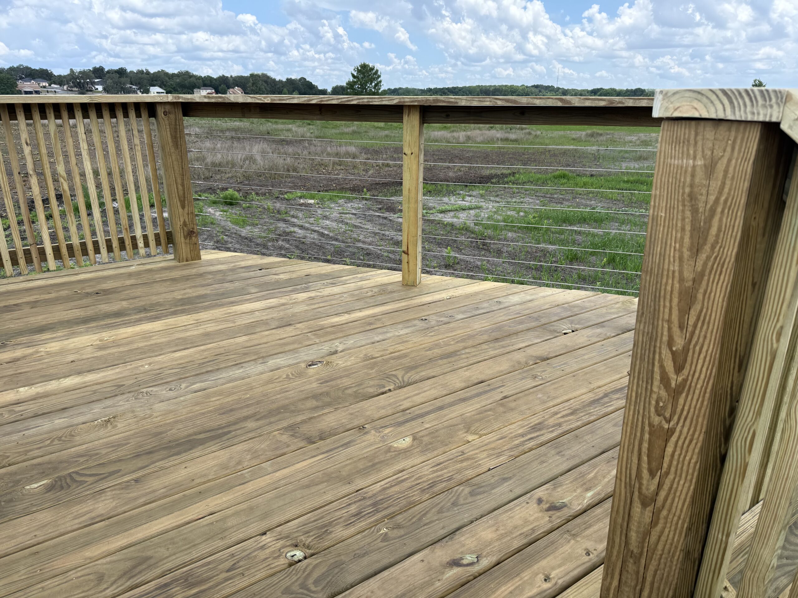 IMG_2568 - JSC Contracting Inc Wooden deck railing with metal cables overlooking a grassy, open field and distant houses under a cloudy sky.