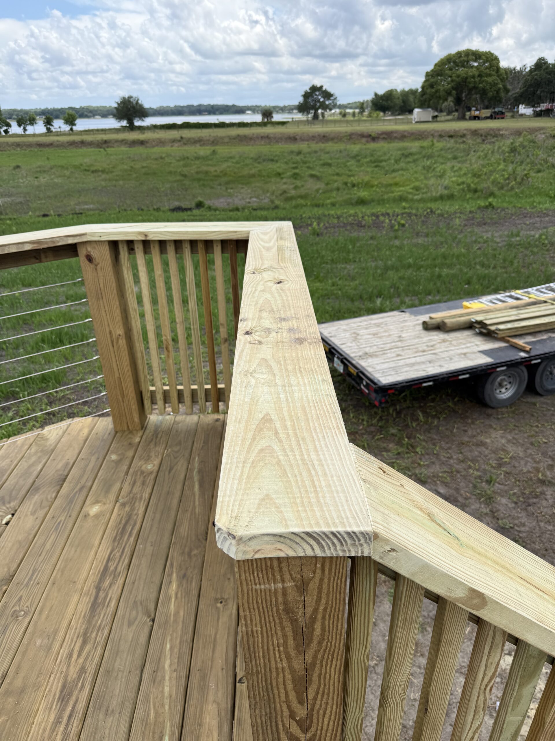 IMG_2569 - JSC Contracting Inc Close-up of a wooden deck railing and top rail with a trailer and building materials in a field beyond.