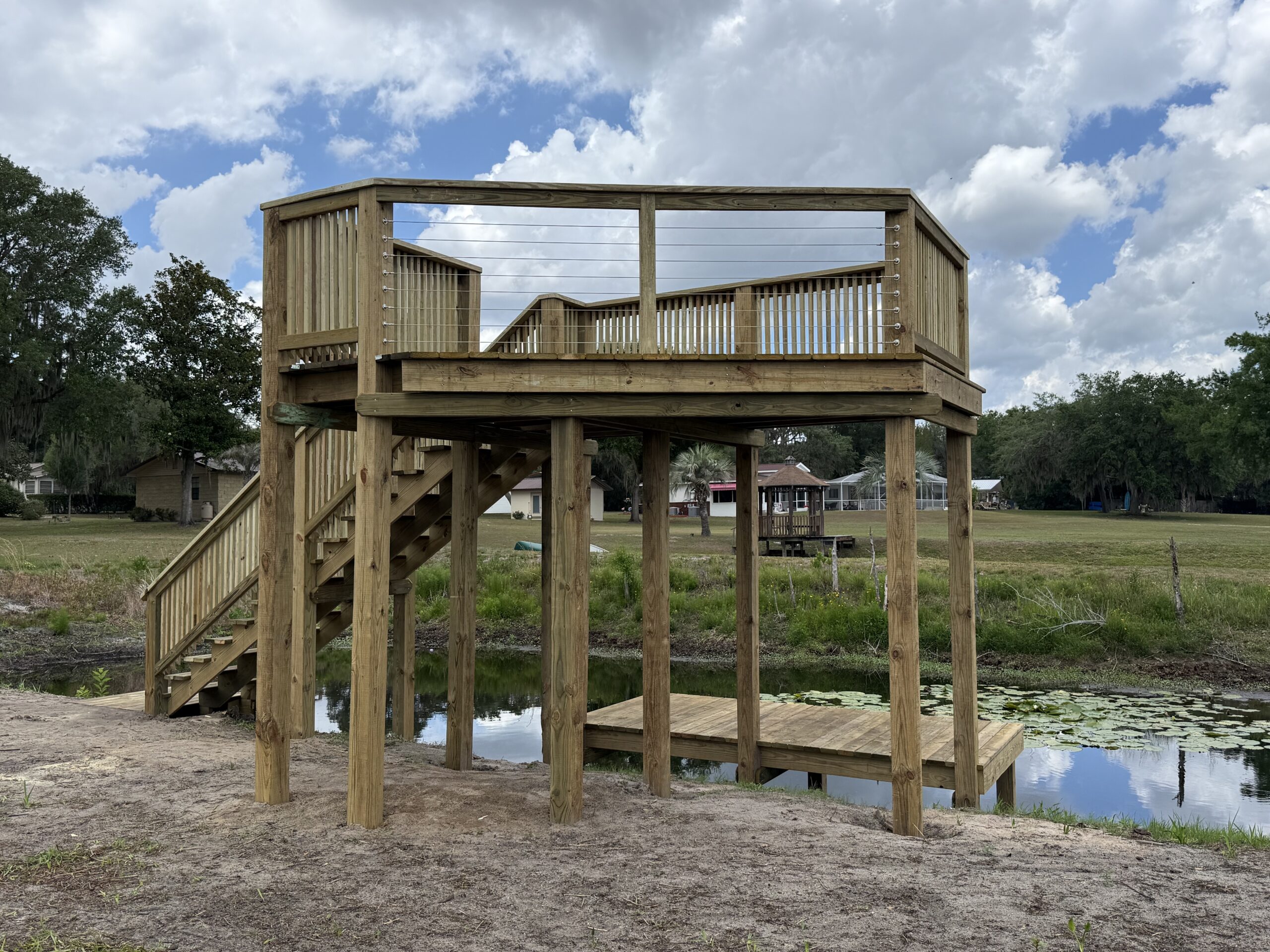 IMG_2578 - JSC Contracting Inc Two-level wooden deck on stilts above a small pond, with stairs on the left and cable railings, in a park with trees and houses in the background under a cloudy sky.