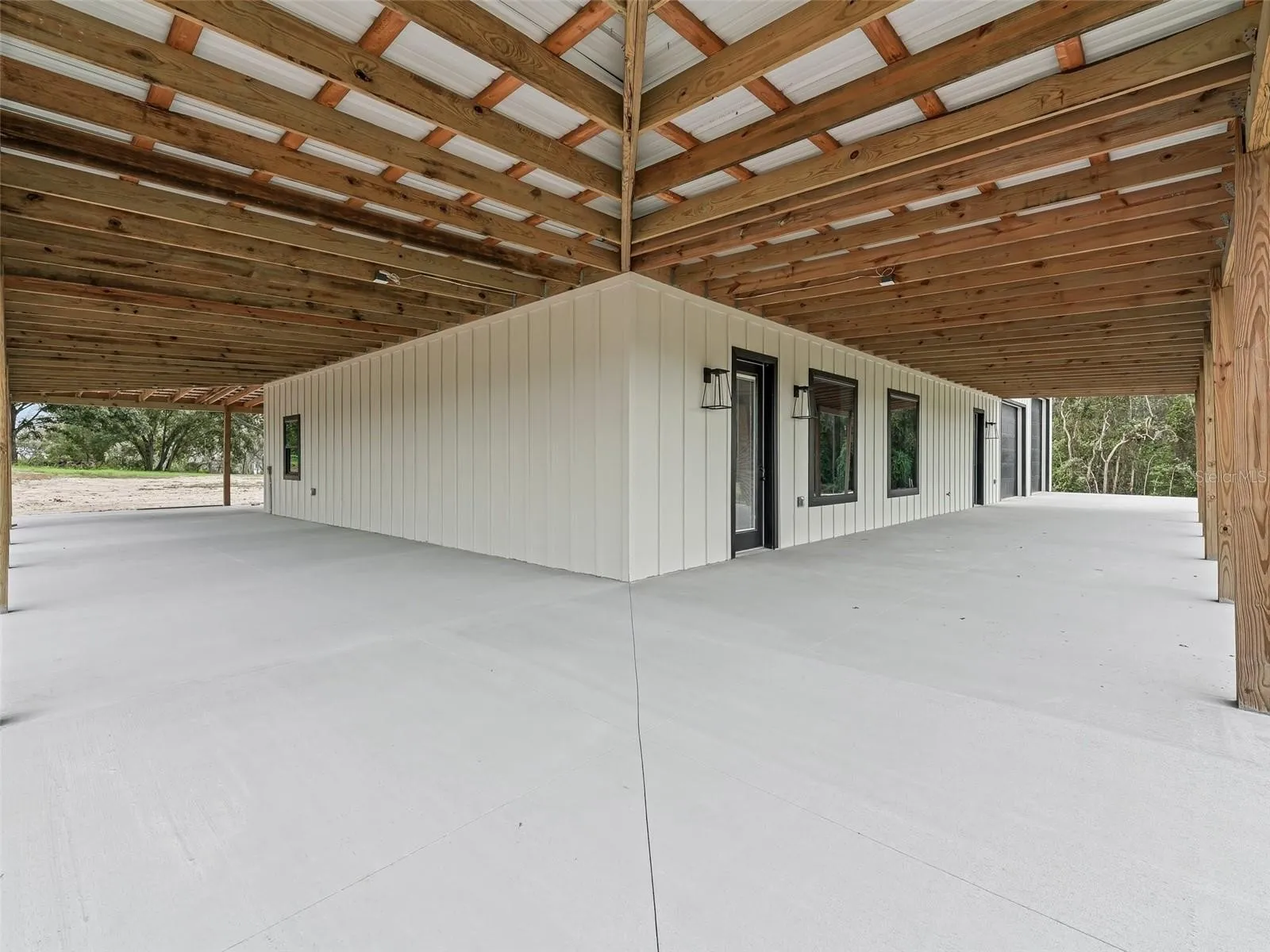 Covered outdoor patio with a white paneled wall, concrete floor, and exposed wooden beams above; exterior landscape visible at the edges.