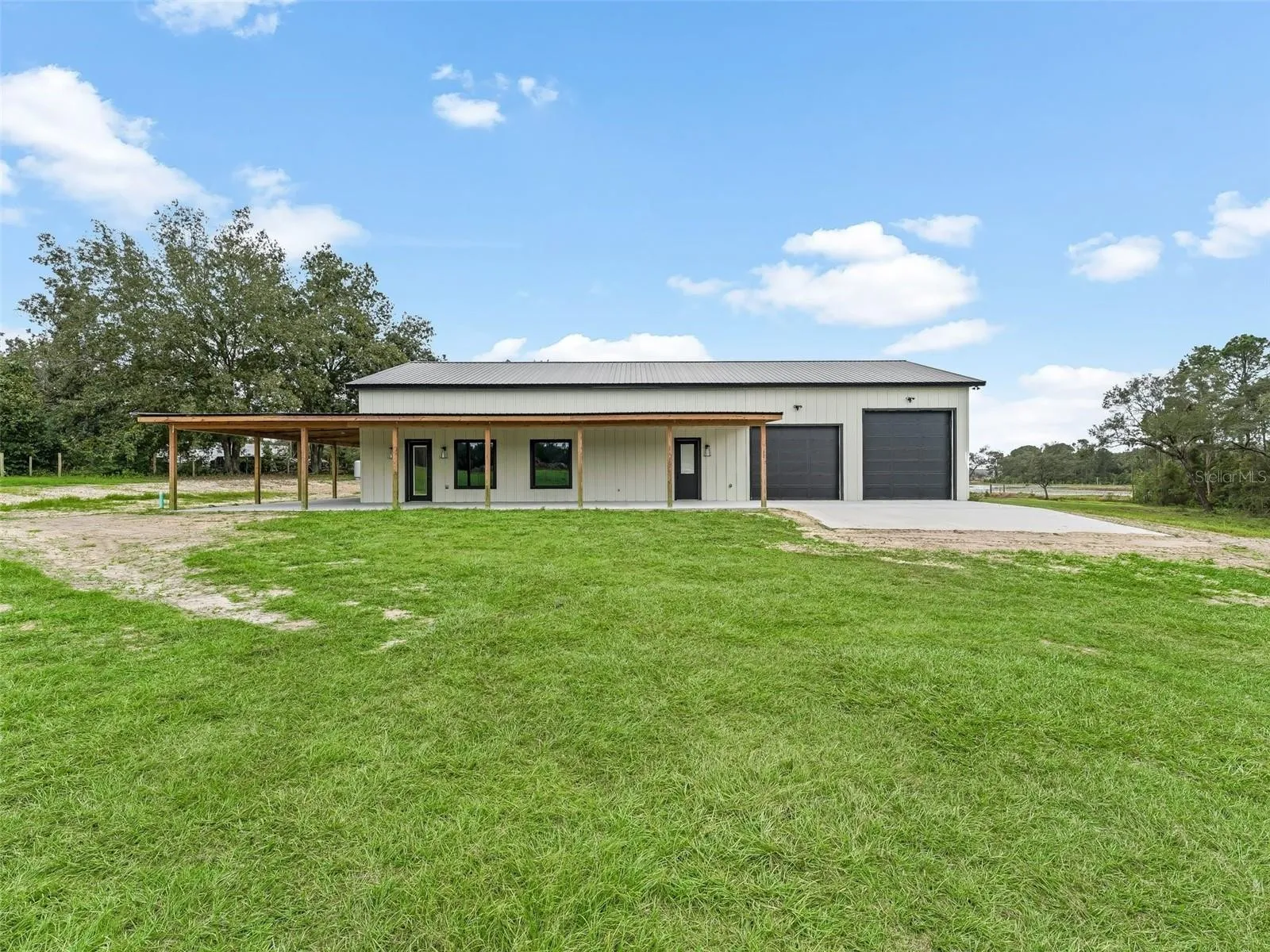 Modern metal building with a wooden carport extending across the front and two large black garage doors, set in a grassy field.