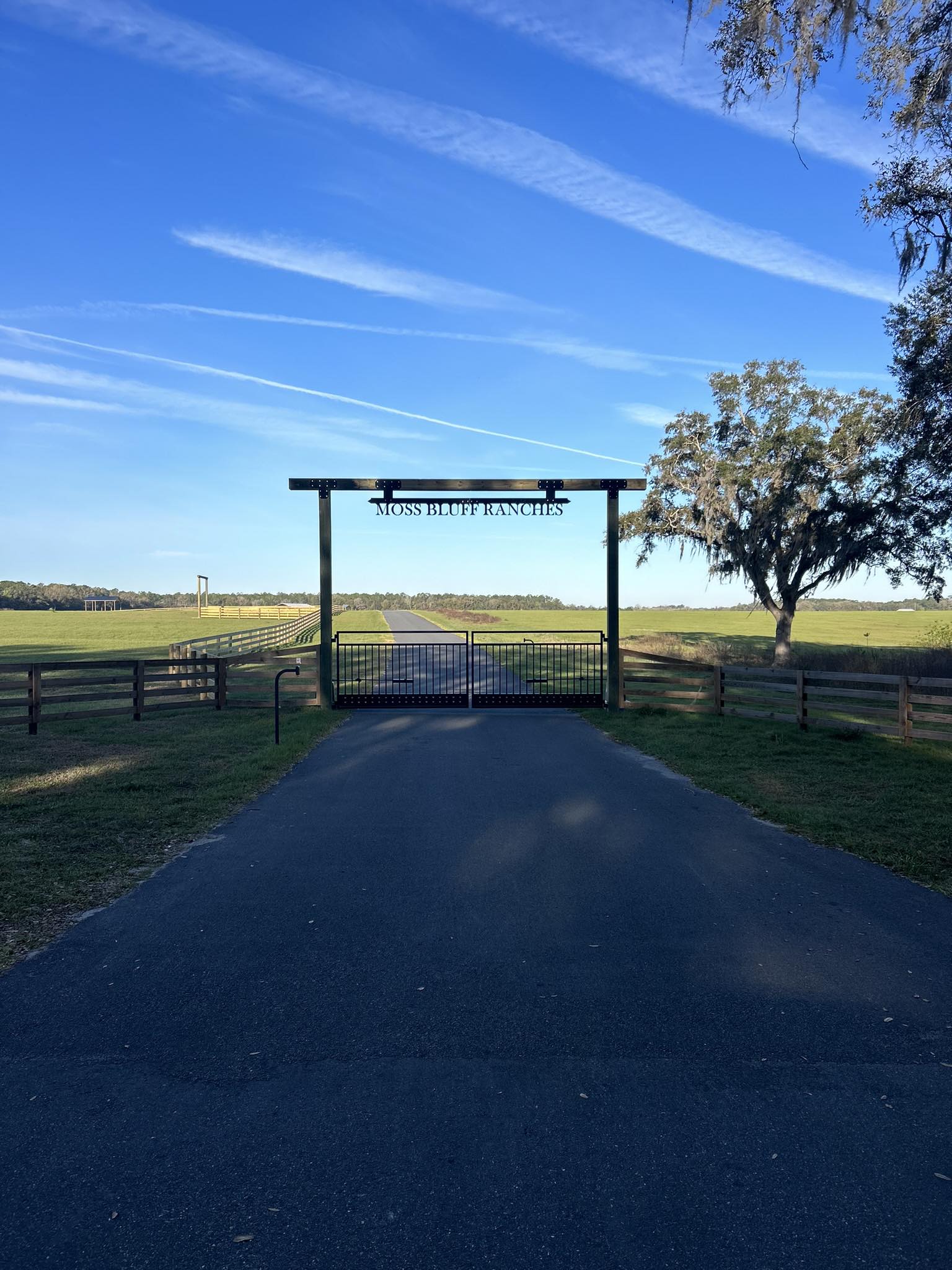 Gated ranch entrance reading Moss Bluff Ranches, with a long paved driveway and wooden fences leading to open fields under a blue sky.