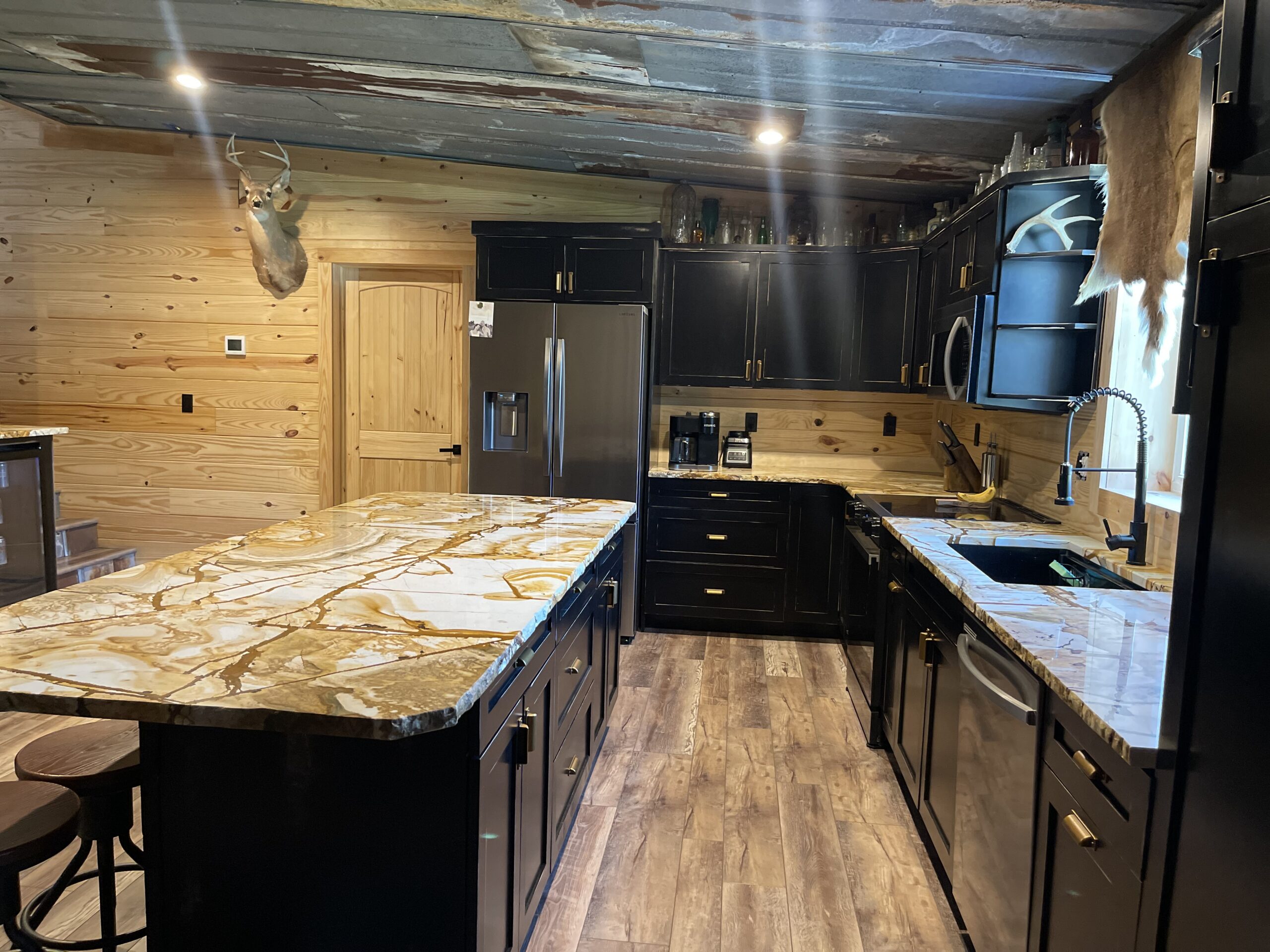 Rustic kitchen with dark cabinetry, marble countertops, a stainless steel refrigerator, and a mounted deer head on a wood-paneled wall.