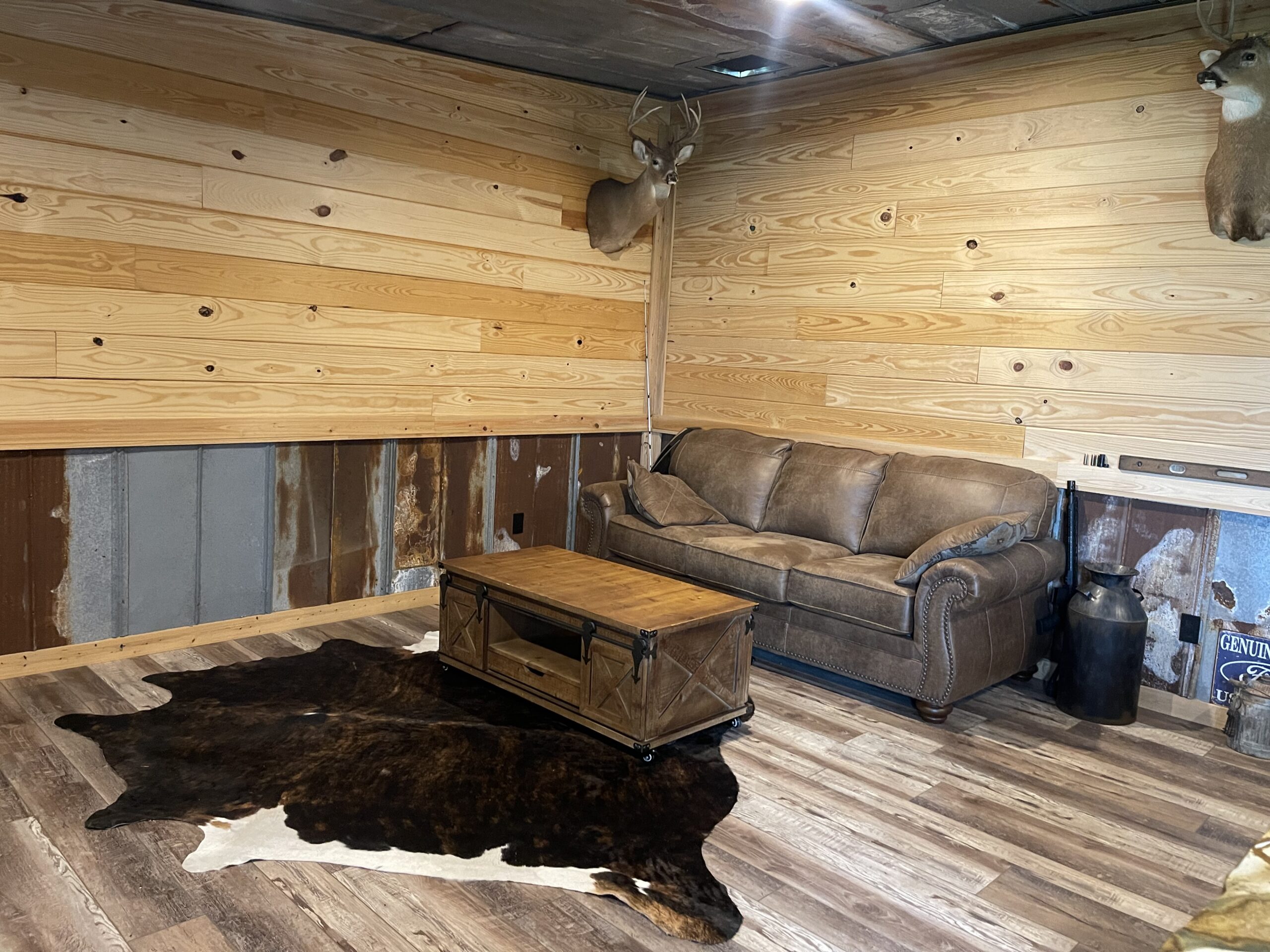 Living room with knotty pine walls, a mounted deer head, brown leather sofa, and a wooden coffee table on a cowhide rug.