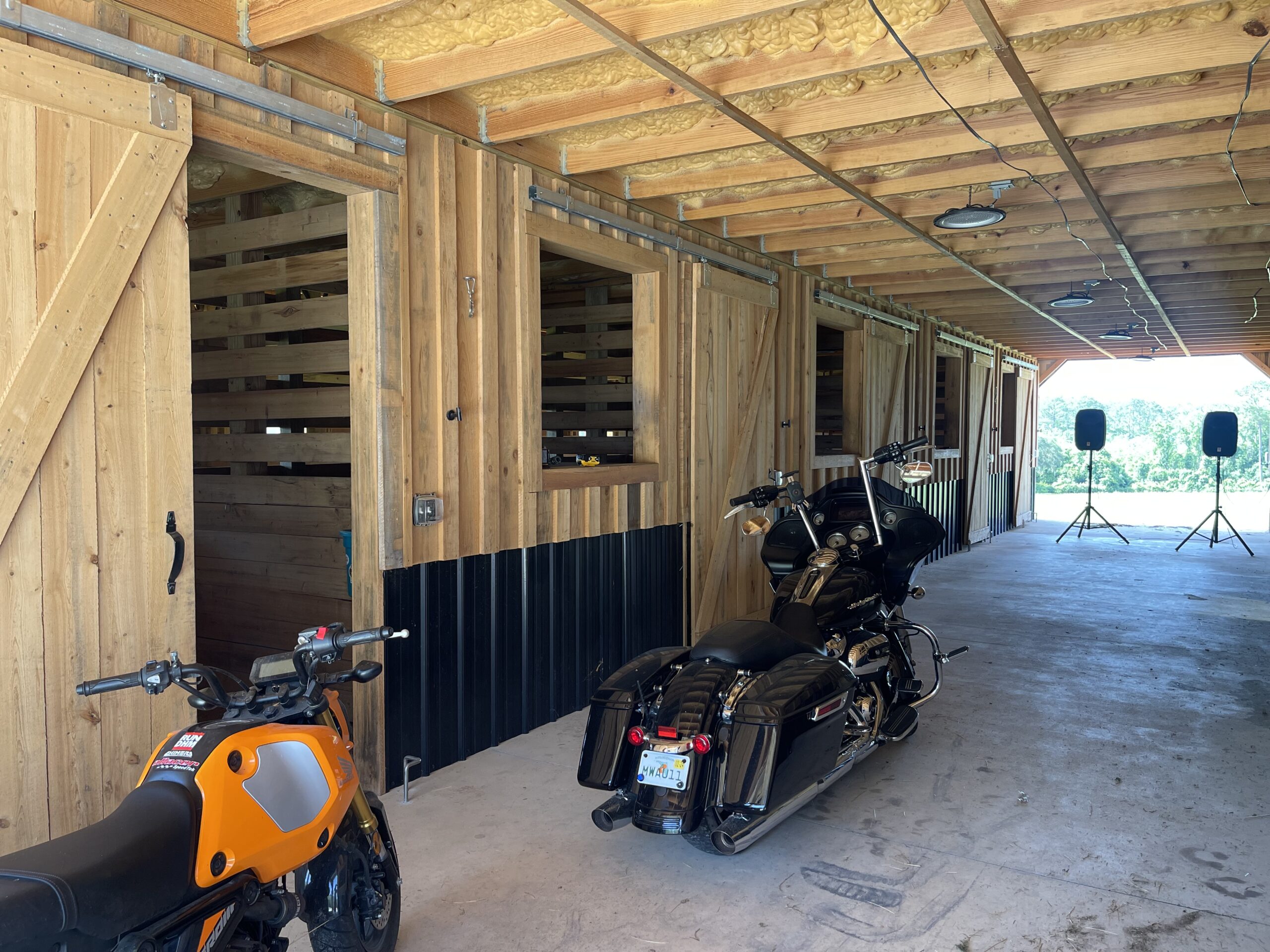 Row of motorcycles parked inside a wooden barn-like garage with exposed ceiling beams and insulation above.