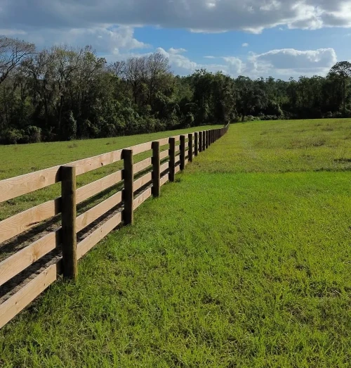 Wooden split-rail fence runs diagonally across a sunlit green field toward a tree line under a blue sky with clouds.