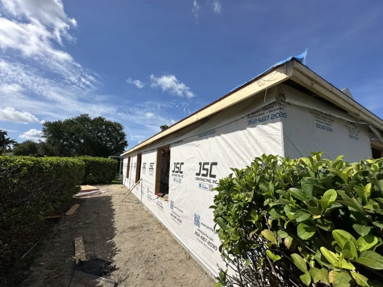 Side view of a single-story house under construction, wrapped in white weather barrier with a clear blue sky above and a hedge along a dirt path